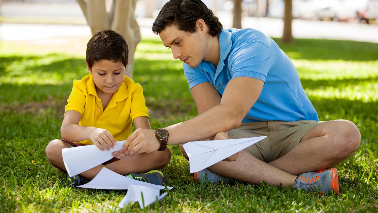 A boy and a man making a paper airplane in a park