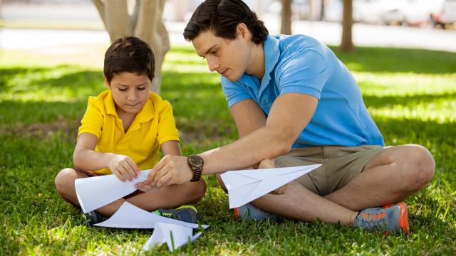 A boy and a man sitting in the grass making paper airplanes 